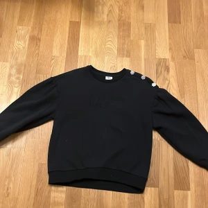 Svart tröja med dekorativa knappar från Zara - Säljer en svart tröja från Zara med texten 'L'amour Réel' på framsidan. Tröjan har dekorativa knappar på axeln som ger en extra touch. Perfekt för en stilren look.❤️