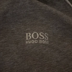 Grå huvtröja från Hugo Boss - Säljer en stilren grå huvtröja från Hugo Boss med dragkedja och broderad logga på bröstet. Tröjan har långa ärmar och en bekväm passform, perfekt för en avslappnad look.
