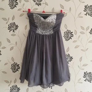 Grå axelbandslös klänning från Sisters Point, Strl:34/XS - Snygg grå axelbandslös klänning från Sisters med glittrig paljettöverdel. Kjolen är luftig och faller fint. Perfekt för en festlig kväll!