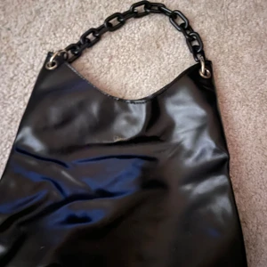 Svart axelväska med kedjehandtag - Snygg svart axelväska i syntetmaterial med ett coolt kedjehandtag. Perfekt för att ge din outfit en edgy touch. Väskan har en minimalistisk design och är lätt att matcha med olika stilar.