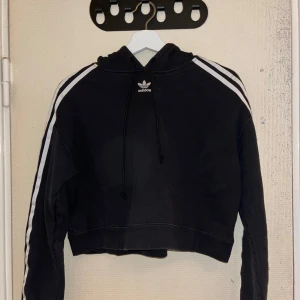 Svart croppad hoodie från Adidas - Säljer en svart croppad hoodie från Adidas med de klassiska vita ränderna längs ärmarna. Hoodien har en justerbar huva och är perfekt för en sportig look. Den är både bekväm och stilren.