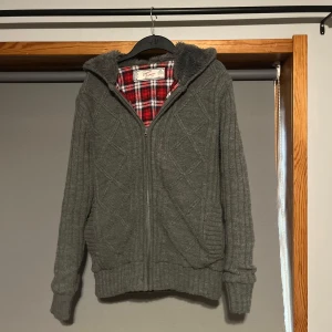 Cardigan jacka med foder och päls - Mysig grå stickad kofta med dragkedja och huva. Insidan är fodrad med röd och vit rutig flanell, vilket ger extra värme och stil. Perfekt för kyliga dagar.