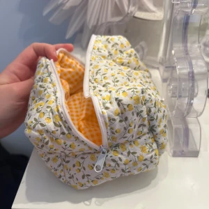 Blommig necessär med dragkedja - Charmig necessär med blommigt mönster i gult och grönt på vit bakgrund. Insidan har ett orange och vitt rutigt mönster. Perfekt för att förvara smink eller andra småsaker. Stängs med en dragkedja.