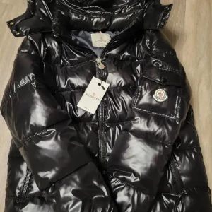Svart pufferjacka från Moncler - Snygg svart pufferjacka från Moncler med glansig finish och dragkedja. Jackan har en hög krage och praktiska fickor. Perfekt för kyliga dagar. Pris går att diskutera! NFC-scan funkar!