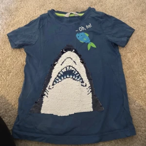 Blå t-shirt med hajmotiv - Säljer en blå t-shirt med ett coolt hajmotiv och texten 'Oh, no!'. Perfekt för den som gillar havstema. T-shirten har korta ärmar och är gjord i ett mjukt material.