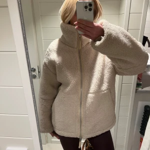 Beige teddyjacka med dragkedja - Säljer denna mysiga beige teddyjacka med dragkedja och hög krage. Perfekt för kyliga dagar med sin fluffiga och varma design. Jackan har en avslappnad passform och långa ärmar.
