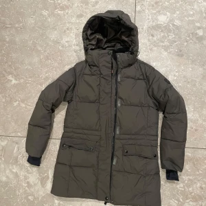 Mörkgrön vadderad jacka med huva - Säljer en  mörkgrön vadderad med huva. Jackan har dragkedja och knappar framtill samt praktiska fickor. Perfekt för kyliga dagar.