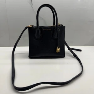 Svart handväska från Michael Kors - Snygg svart handväska från Michael Kors i skinn med gulddetaljer. Väskan har både handtag och en justerbar axelrem för flexibel användning. Perfekt för att bära dina nödvändigheter med stil.