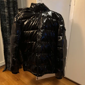 Moncler Maya  - Snygg svart glansig dunjacka från Moncler med dragkedja och hög krage. Jackan har ett klassiskt Moncler-märke på ärmen och är perfekt skick!