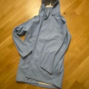 Ljusblå regnjacka med huva - Snygg ljusblå regnjacka med huva och knappar. Perfekt för regniga dagar med sin långa ärm och stilrena design. Jackan har en praktisk passform och är ett måste för garderoben.