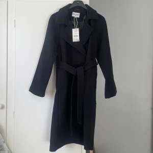 Svart kappa med knytskärp - En svart lång kappa från NA-KD med knappstängning och bälte/knytskärp i midjan. Helt ny, endast provad. Nypris 999kr. • Istoppsfickor i sidorna • Sprund/slits bak • Längd: cirka 105 cm från axeln