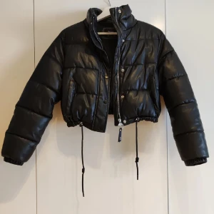 Svart pufferjacka - Snygg svart pufferjacka med croppad design och dragkedja. Jackan har långa ärmar och snörning i nederkant för justerbar passform. Perfekt för kyligare dagar. Säljer då den är för liten. Pris kan diskuteras.