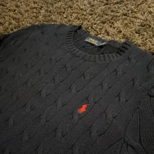 Mörkblå stickad tröja från Polo Ralph Lauren - Säljer en snygg mörkblå stickad tröja från Polo Ralph Lauren. Den har ett klassiskt kabelstickat mönster och den ikoniska röda loggan på bröstet. Perfekt för både höst och vinter, med långa ärmar och ribbade muddar. Passar både till vardags och lite finare tillfällen. Nypris 1400kr