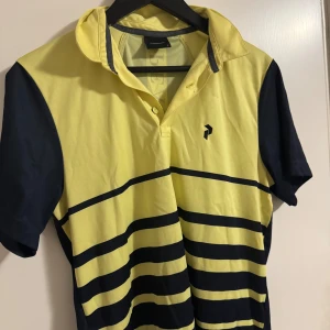 Gul och marinblå randig pikétröja - Snygg gul och marinblå randig pikétröja från Peak Performance. Tröjan har korta ärmar och en klassisk krage med knappar. Perfekt för en sportig look eller en avslappnad dag. Storlek S men passar även M
