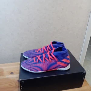 Färgglada fotbollsskor med snörning - Säljer ett par färgglada fotbollsskor med snörning i blått och rosa. Skorna har en modern design med en mönstrad ovandel och vit sula.