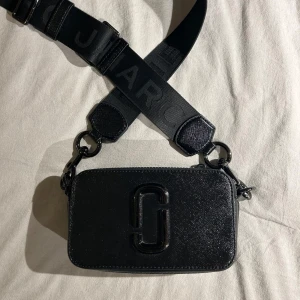 Svart axelväska från Marc Jacobs - Snygg svart väska från Marc Jacobs med justerbar rem. Säljer då den inte kommer till andvändning längre. Jätte fint skick men 2 flärpar på dragkedjan har tyvärr lossnat. Ny pris 4315.