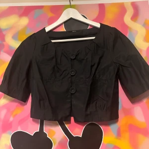 Svart bolero från Comme Ça - Snygg svart bolero från Comme Ça med trekvartsärm och stora knappar framtill. Perfekt för en stilren look.