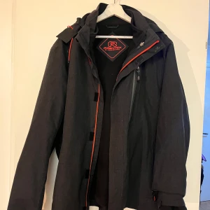Svart regnjacka från Raintex - Snygg svart regnjacka från Raintex med röda detaljer. Jackan har en praktisk dragkedja och flera fickor. Perfekt för alla väder med sin funktionella design.