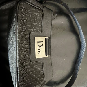 Svart axelväska från Dior - Snygg svart axelväska från Dior med monogrammönster och en elegant metallplatta med logotyp. Väskan har en praktisk dragkedja och justerbar axelrem. Perfekt för att ge en lyxig touch till din outfit.
