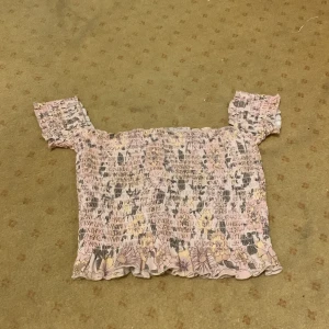 Blommig offshoulder topp från BikBok - Söt offshoulder topp från BikBok i rosa med blommigt mönster. Toppen har en smockad design och volangdetaljer vid ärmslut och nederkant. Perfekt för en somrig look!