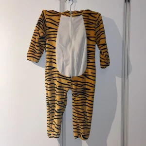 Tigerdress och tofflor - Mysig tiger-onepiece för barn i storlek 92 samt medföljande tigertofflor storlek 25/26. Ett par små fläckar förekommer (går eventuellt att tvätta bort). Bra som både utklädningsklädsel och mjukiskläder. 