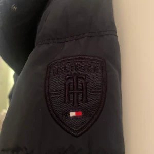 Svart dunjacka från Tommy Hilfiger - Snygg svart dunjacka från Tommy Hilfiger med quiltad design och diskret logotyp på ärmen. Jackan har en praktisk dragkedja och knappar framtill samt en bekväm passform. Perfekt för kyligare dagar.