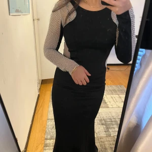 Svart långklänning med pärldetaljer - Elegant svart långklänning med långa ärmar dekorerade med pärlor. Klänningen har en tight passform och meshdetaljer på sidorna som ger en sofistikerad look. Använd 1 gång.