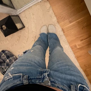 Blå utsvängda lågmidjade jeans med slitningar - Säljer ett par blå lågmidjade utsvängda jeans med slitningar på framsidan och baksidan. De har en klassisk femficksdesign och är i en straight passform. Perfekta för en avslappnad stil. Hålet på baksidan är bara i första tyget!