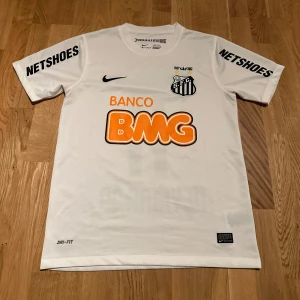  Santos fotbollströja Neymar Jr - Säljer en vit Santos fotbollströja med Neymar Jr och nummer 11 på ryggen. Tröjan har korta ärmar och är tillverkad i Dri-FIT material för optimal komfort. Sponsorer som BMG och Netshoes pryder framsidan och ärmarna. Perfekt för fans av Neymar och Santos FC!