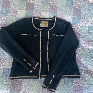 Svart kavaj/kofta med beige detaljer medium L’ecole national - Elegant svart kavaj med kontrasterande beige detaljer och knappar. Perfekt för en stilren look. Långärmad design med dekorationsfickor framtill. En klassiker! Storlek medium.