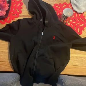 Svart hoodie från Ralph Lauren  - Snygg svart hoodie från Ralph Lauren med dragkedja och huva. Den har en röd broderad logga på bröstet och praktiska fickor framtill. Perfekt för en avslappnad stil. Använd Max 5 gånger då den va för liten mvh 