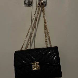 Svart quiltad axelväska med guldkedja - Snygg svart axelväska med quiltat mönster och guldkedja som axelrem. Väskan har en dekorativ guldfärgad låsdetalj framtill. Perfekt för att ge en elegant touch till din outfit.