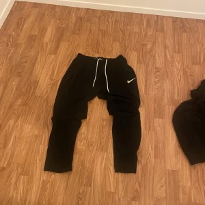 Svarta mjukisbyxor från Nike - Säljer ett par svarta mjukisbyxor från Nike med vit snörning i midjan och den klassiska Nike-loggan på benet. Perfekta för en avslappnad stil eller träning.