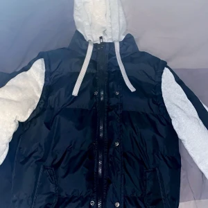 Svart pufferjacka från Blend - Snygg svart pufferjacka från Blend med grå huva och ärmar. Jackan har dragkedja och knappar framtill, perfekt för kyliga dagar. Den har en casual stil med en bekväm passform.