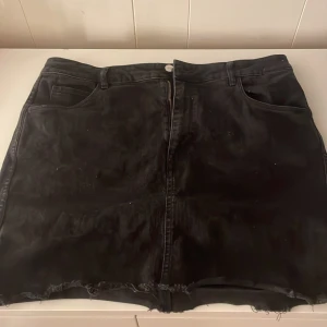 Svart jeanskjol från &Denim - Snygg svart jeanskjol från &Denim i storlek EUR 44. Kjolen har en klassisk femficksdesign med rå kant nertill för en trendig look. Perfekt för en avslappnad stil.