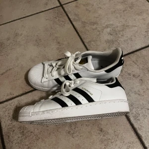 Adidas Superstar sneakers - Klassiska vita Adidas Superstar sneakers med svarta ränder och snörning. Perfekta för en stilren och sportig look.
