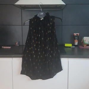 Svart blus med mönster från H&M - Säljer en elegant svart blus från H&M med ett subtilt mönster i guld. Blusen har en dekorativ krage med knytband och är ärmlös, vilket gör den perfekt för både vardag och festliga tillfällen.