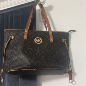 Brun shoppingväska från Michael kors  - Snygg brun shoppingväska från Michel kors  med ett elegant rutigt mönster och gyllene detaljer. Väskan har långa handtag och en dekorativ logotyp i guld på framsidan. Perfekt för att bära med sig allt man behöver under dagen.