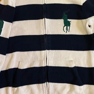 Randig kofta från Ralph Lauren - Snygg randig kofta från Ralph Lauren i marinblått och vitt. Koftan har en dragkedja framtill och två fickor. Den är dekorerad med det klassiska gröna hästlogotypen på bröstet.
