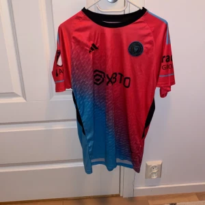 Röd och blå Inter Miami fotbollströja från Adidas - Snygg Inter Miami fotbollströja från Adidas i röd och blå färg med Messi 10 på ryggen. Tröjan har korta ärmar och är tillverkad i Aeroready-material för optimal komfort. Perfekt för fotbollsfans!