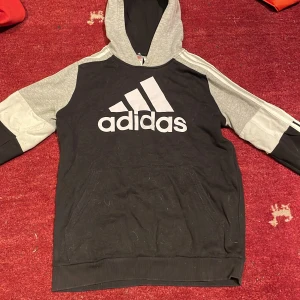 Svart och grå hoodie från Adidas - Säljer en svart och grå hoodie från Adidas med den klassiska loggan på bröstet. Tröjan har en huva och långa ärmar med de ikoniska tre ränderna längs ärmarna. Perfekt för en sportig look eller avslappnad stil.