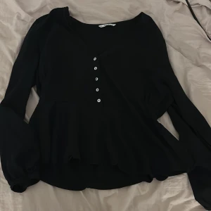 Svart blus från ONLY - Säljer en elegant svart blus från ONLY med v-ringning och knappar framtill. Blusen har långa ärmar och en lätt volangdetalj vid nederkanten. Perfekt för en stilren look.