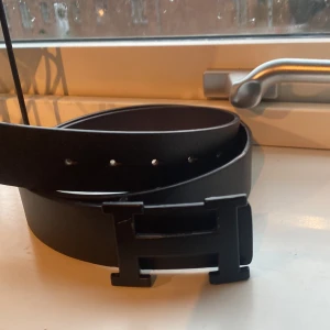 Svart bälte med H-spänne - Snyggt svart bälte med ett stilrent H-format spänne. Perfekt för att ge en elegant touch till din outfit. Bältet är gjort av skinn och har flera hål för justering.