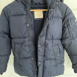 Mörkblå dunjacka från Zara Kids - Säljer en mörkblå dunjacka från Zara Kids. Jackan har en mysig huva och stängs med både dragkedja och knappar. Perfekt för kalla dagar med sin vadderade design. Dock en liten värmeskada på ärmen, se foto