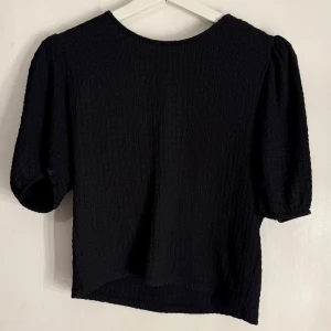 Svart blus med puffärmar - Snygg svart blus med puffiga trekvartsärmar och en rund halsringning. Tyget har en texturerad yta som ger en elegant touch. Perfekt för att klä upp eller ner beroende på tillfälle. 