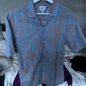 Blå och orange skjorta från H&M - Snygg kortärmad skjorta från H&M i en blå och orange färgkombination. Skjortan har ett abstrakt mönster och knappar framtill. Perfekt för en avslappnad stil.