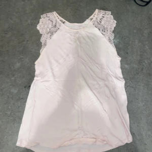 Ljusrosa ärmlös topp med spetsdetaljer - Säljer en söt ljusrosa ärmlös topp med vackra spetsdetaljer på axlarna. Perfekt för varma dagar och kan enkelt matchas med olika outfits.