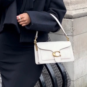 Vit axelväska från Coach - Snygg vit axelväska från Coach med gulddetaljer och ett stort C-spänne på framsidan. Väskan har en elegant design och är perfekt för att lyfta vilken outfit som helst. Den har en justerbar axelrem och är tillverkad i skinn.