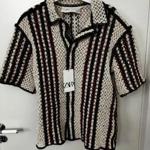 Zara stickad overshirt strl L - Säljer en unik stickad tröja från Zara med korta ärmar och ett randigt mönster i beige, svart och vinrött. Perfekt för en stilren look med en twist. Tröjan har en klassisk krage och är i storlek L. Ej använd i nyskick