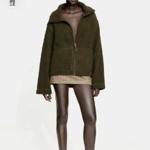 Olivgrön teddyjacka med dragkedja - Mysig olivgrön teddyjacka med dragkedja framtill. Jackan har en hög krage och långa ärmar, perfekt för kyligare dagar.